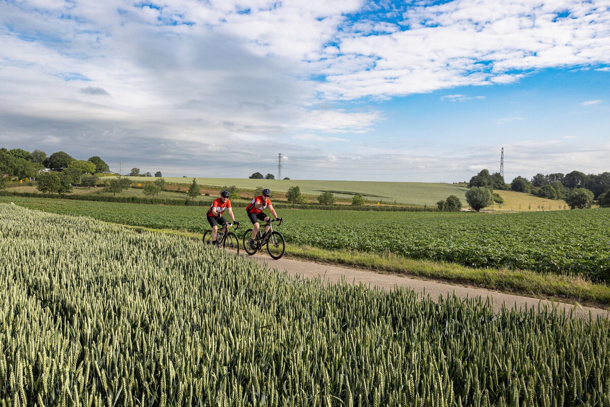 Vebego Limburgs Mooiste - twee wielrenners in Zuid-Limburgse landschap doen mee aan de wedstrijd. Op de voor- en achtergrond zie je velden en een blauwe lucht.