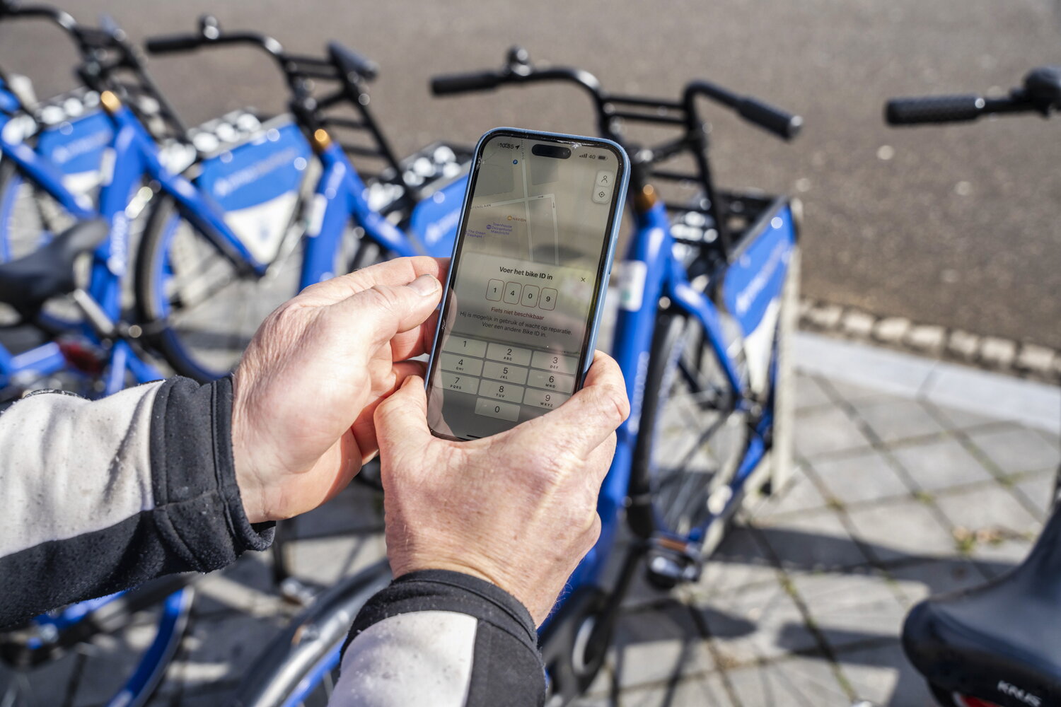 Persoon heeft telefoon in zijn handen om een blauwe deelfiets van Arriva te reserveren of openen. De deelfietsen staan naar elkaar in een rek.