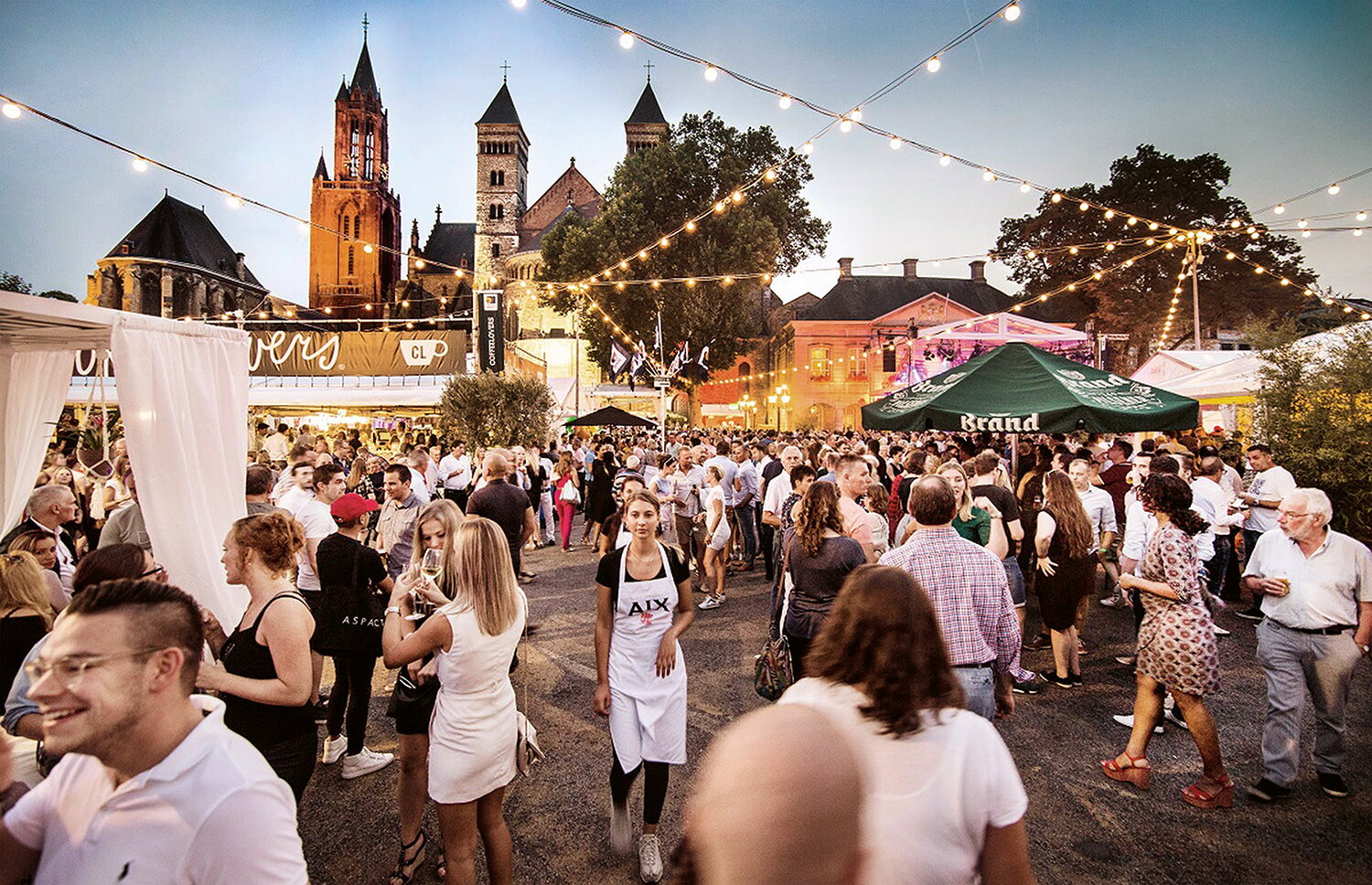 Sfeerbeeld Preuvenemint Vrijthof Maastricht met bezoekers die met elkaar staan te praten in groepjes. Tenten in wit en groen waar je eten kunt bestellen. Lampjes die getrokken zijn om sfeer te geven nu de schering valt. Op de achtergrond zie je de St. Servaaskerk die is verlicht vanuit onderen.