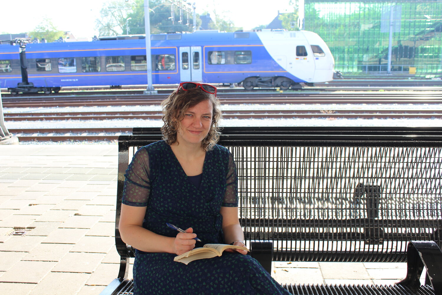 Marianne zit op een bankje op station Maastricht en maakt een sudoku. Achter haar een blauwe trein van Arriva