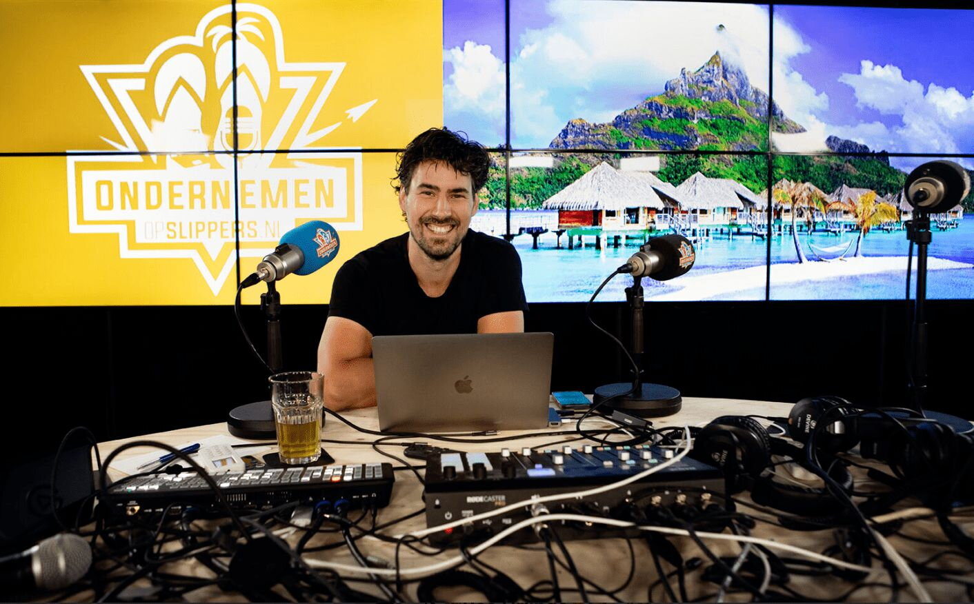Bas Urlings zit in een studio met een laptop en techniek voor zich. Op de achtergrond het logo van Ondernemer op slippers en een foto van rieten huisjes in zee