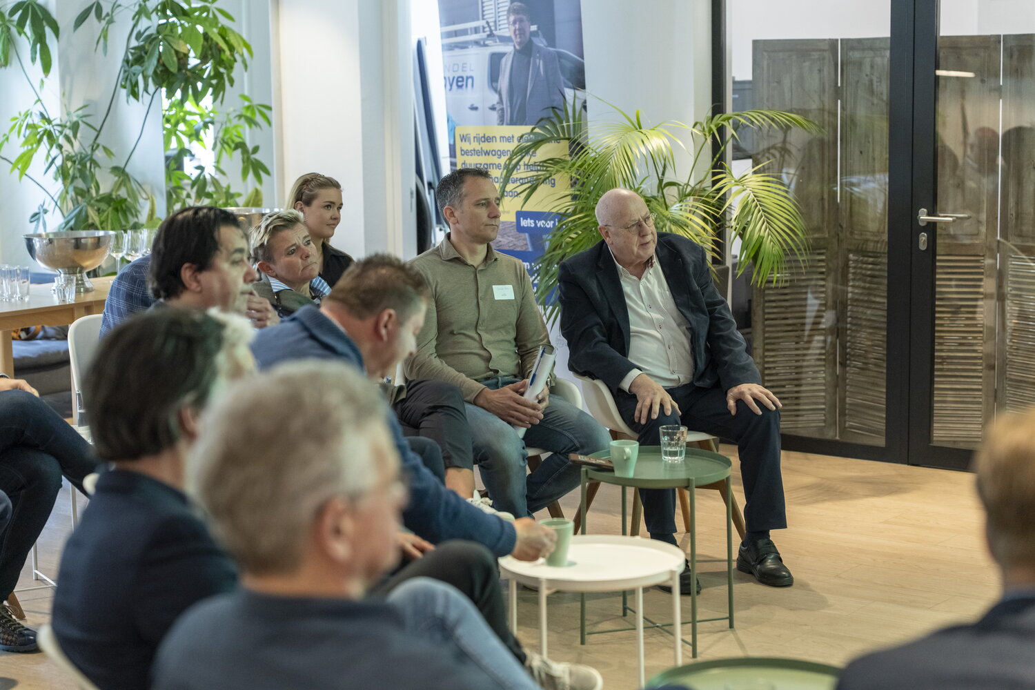 Groep mensen zit op stoelen te luisteren en twee lege tafeltjes