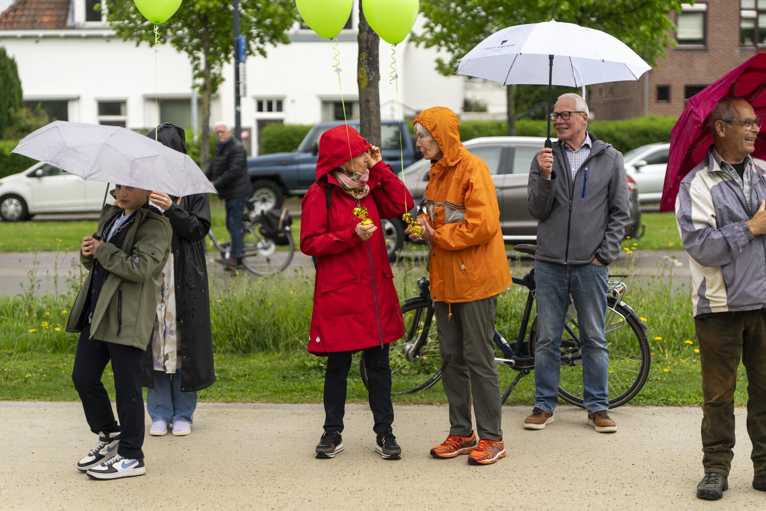 Mensen staan in de regen met paraplu's en regenjassen. Twee dames in een rode en oranje jas praten met elkaar